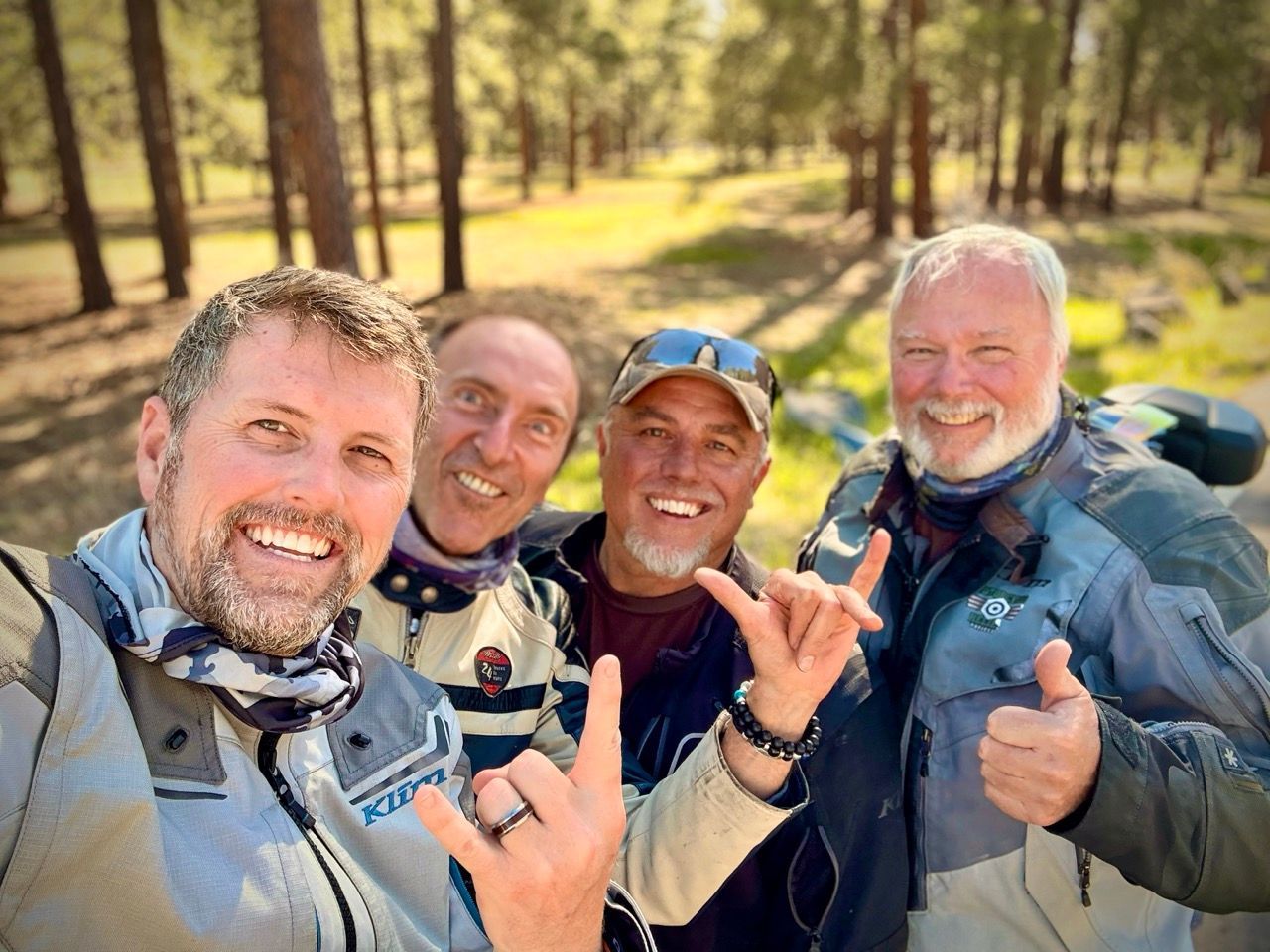 Four men in motorcycle gear smile for a selfie outdoors. Forest background.