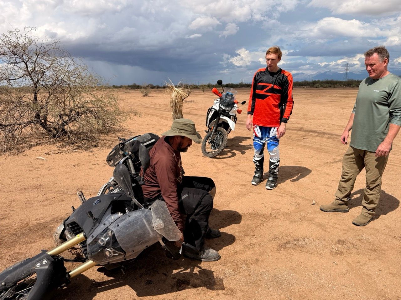 Man beside a fallen motorcycle being helped by two others in a desert landscape with storm clouds.