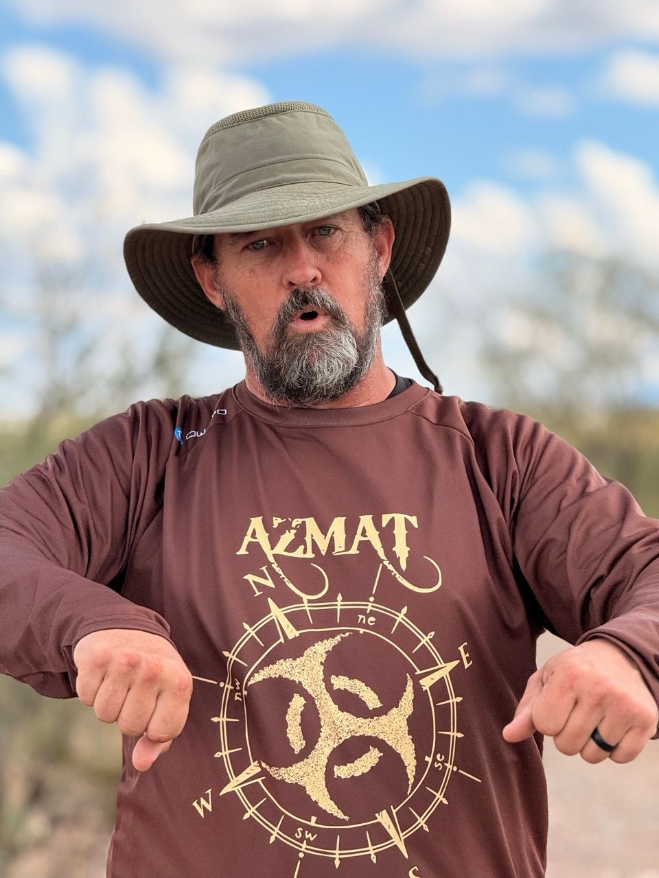 Man in brown hat and shirt pointing down, with a compass-like graphic on shirt; outdoors.