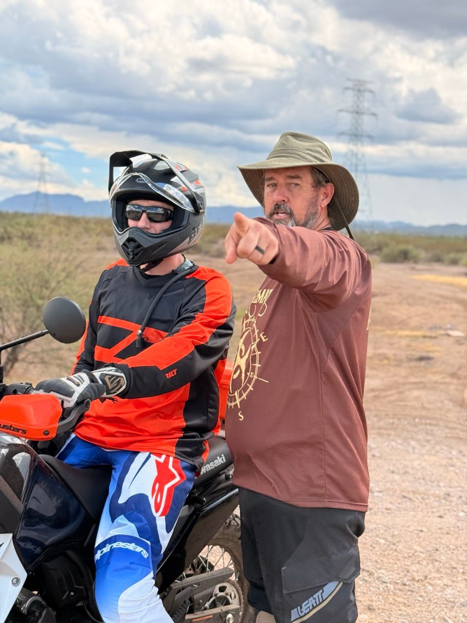 Man on dirt bike and man in hat pointing, desert background.