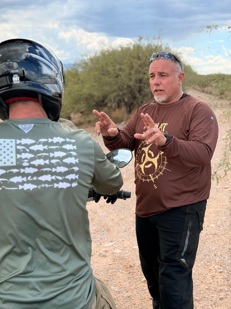 Man in brown shirt gesturing to person on motorcycle in a desert setting.