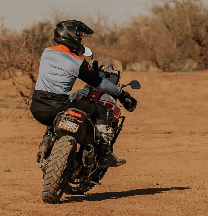 Person riding a motorcycle on dirt road, a passenger behind. Both wearing helmets and riding gear in a desert setting.