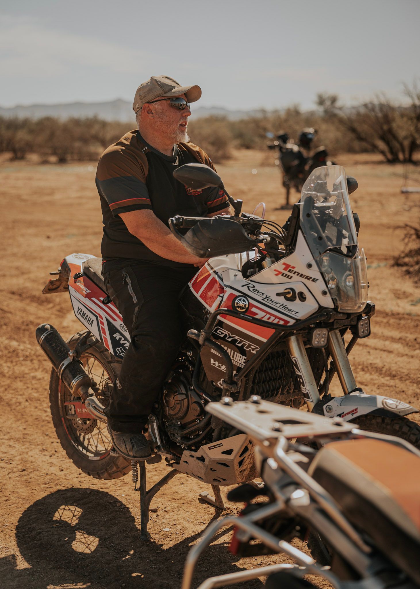 Man on a motorcycle in desert terrain; wearing a hat and protective gear.