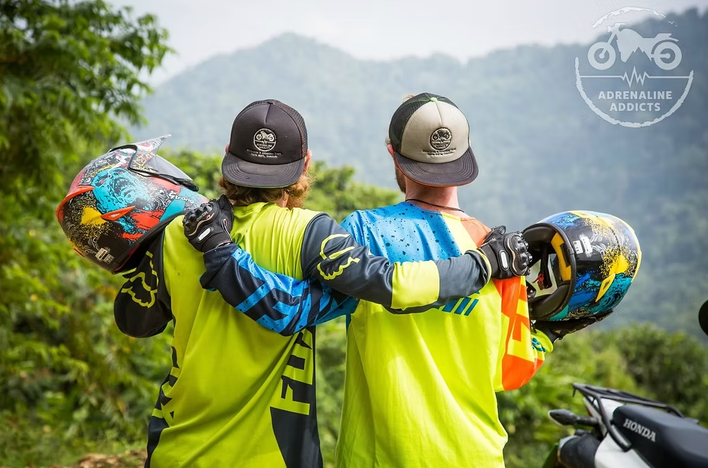 Two people in motocross gear, arms around each other, helmets in hand, overlooking a mountainous landscape.