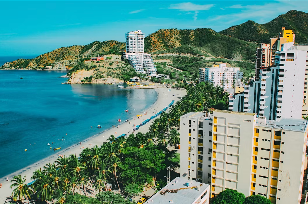 Coastal city with a white sand beach, turquoise water, palm trees, and high-rise buildings against a mountain backdrop.