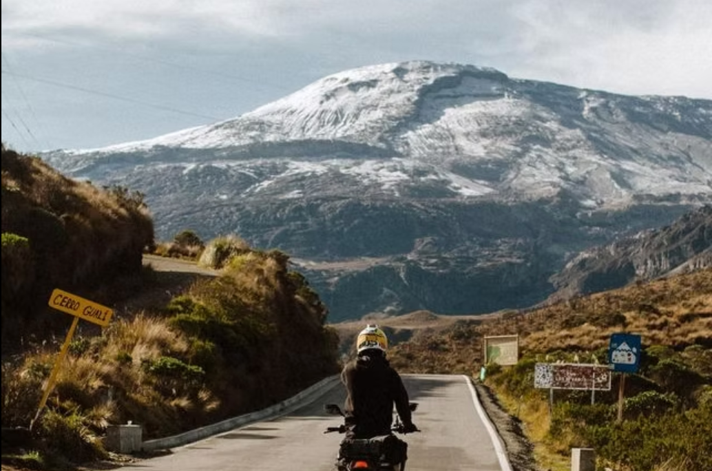 Motorcyclist on a road with a snowy mountain in the background. Roadside signs are visible.