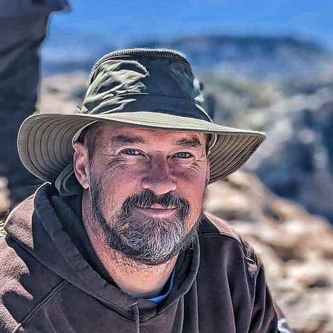 Man with graying beard and hat smiles outdoors.