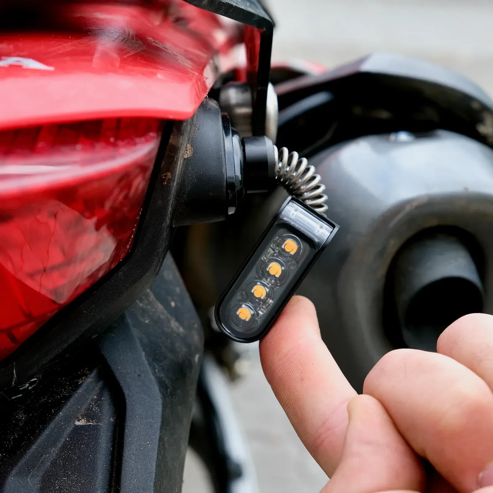 Motorcycle turn signal light with orange LEDs, being held by a finger, attached to a red fender.