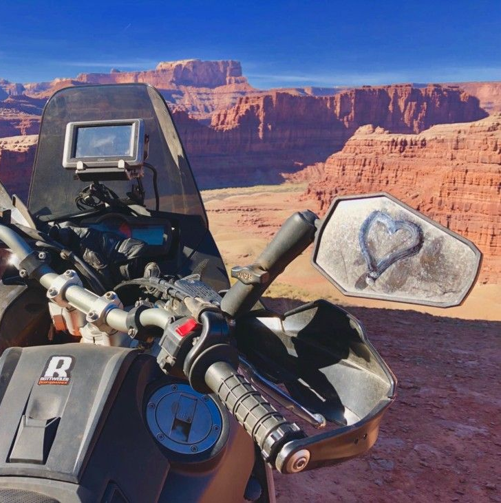 Motorcycle cockpit with a desert canyon backdrop. Handlebars, mirror with a heart drawn in the dirt, and a screen visible.