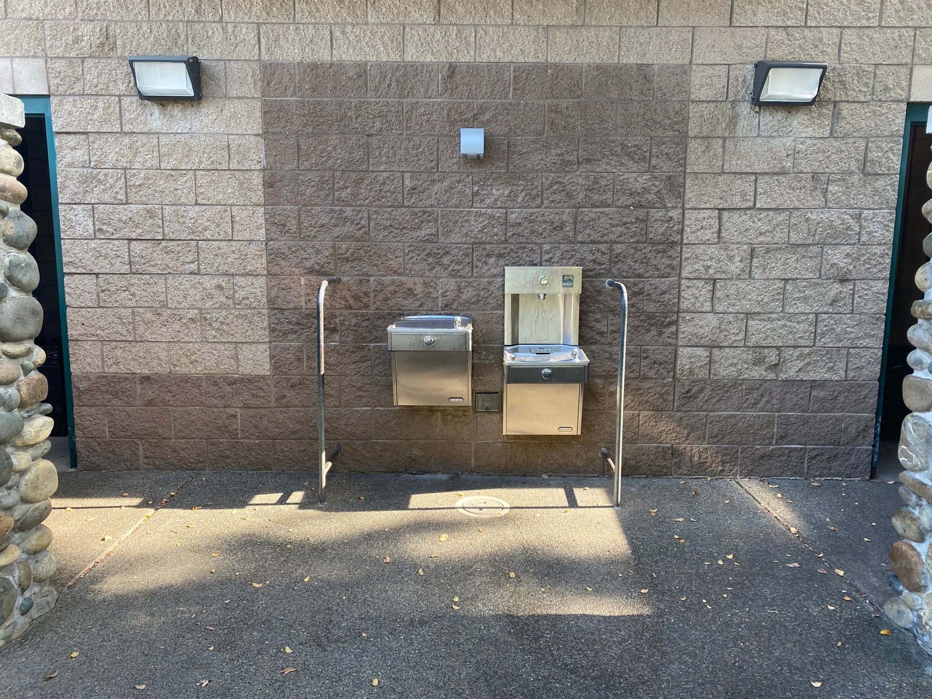 Outdoor public drinking fountain with two stainless-steel water dispensers set in a stone wall.