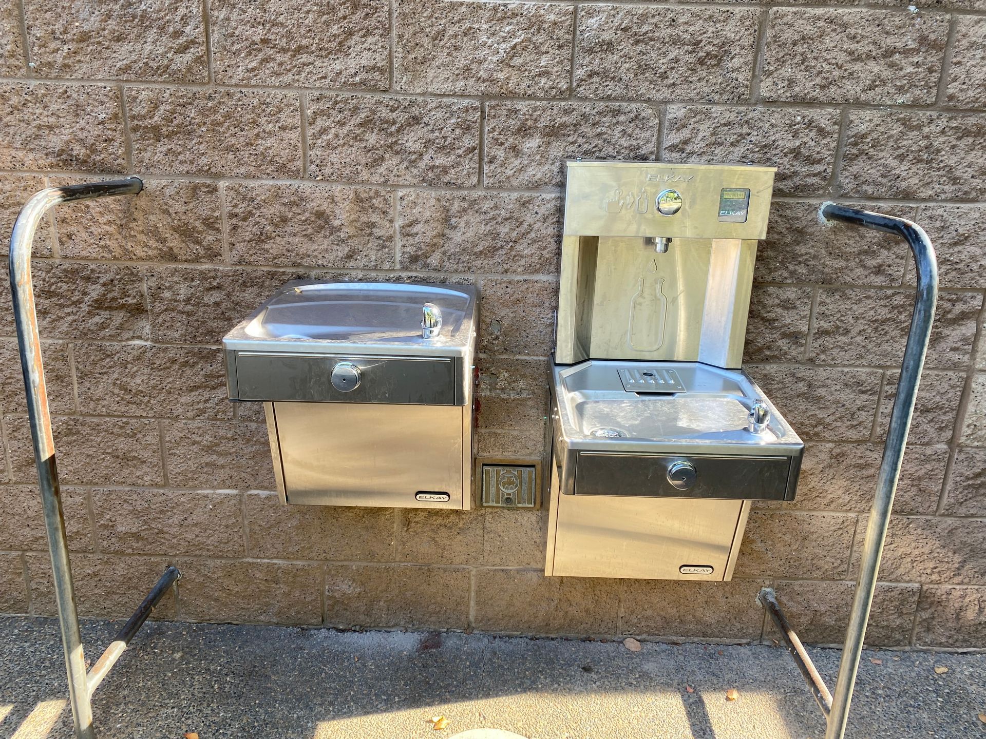 Two stainless steel drinking fountains mounted on a stone wall, with grab bars on both sides.