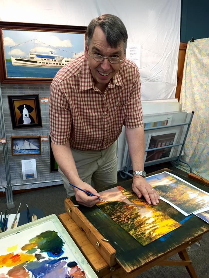 Man painting a watercolor at a table. He wears glasses and a blue shirt, working in a well-lit room.
