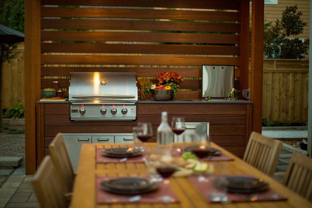 Outdoor kitchen with grill, built-in cabinets, wooden privacy screen, and dining table set for dinner.