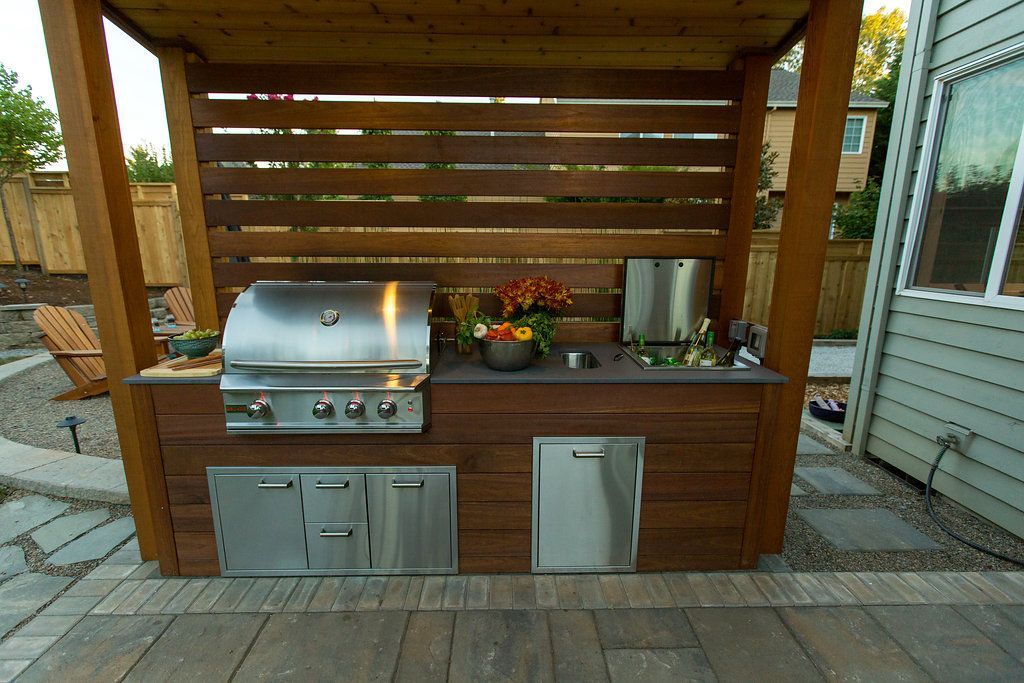 Outdoor grilling station with a grill, fridge, and storage, under a wooden pergola.