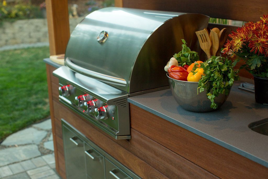 Outdoor kitchen with stainless steel grill, bowl of vegetables, and wooden cabinetry.