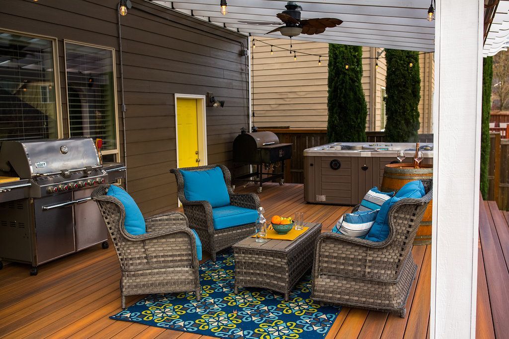 Outdoor deck with seating, grill, and hot tub.  Blue cushions, wood deck, white ceiling, and string lights.