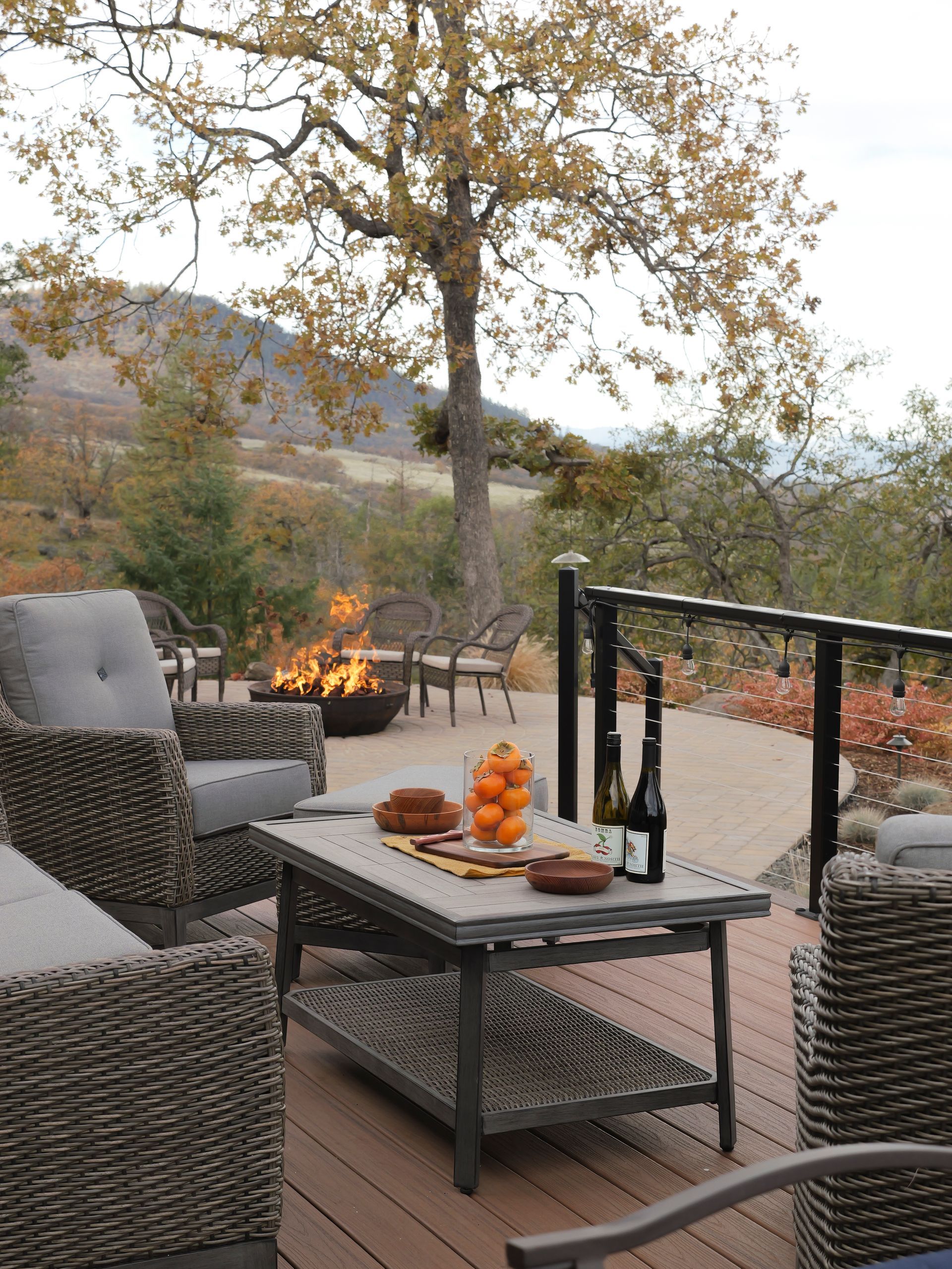 Outdoor deck with wicker furniture, fire pit, fall foliage, and wine bottles.