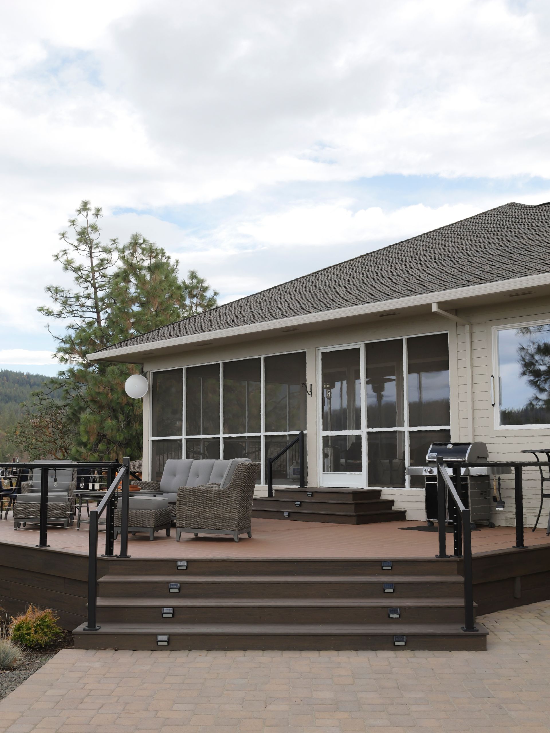 Wooden deck with steps leading to a screened porch with outdoor furniture, overlooking a stone patio and yard.
