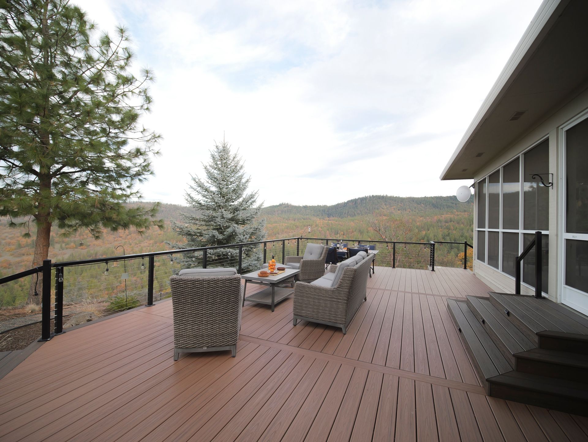 Wooden deck with outdoor furniture, overlooking a forested valley under a cloudy sky.