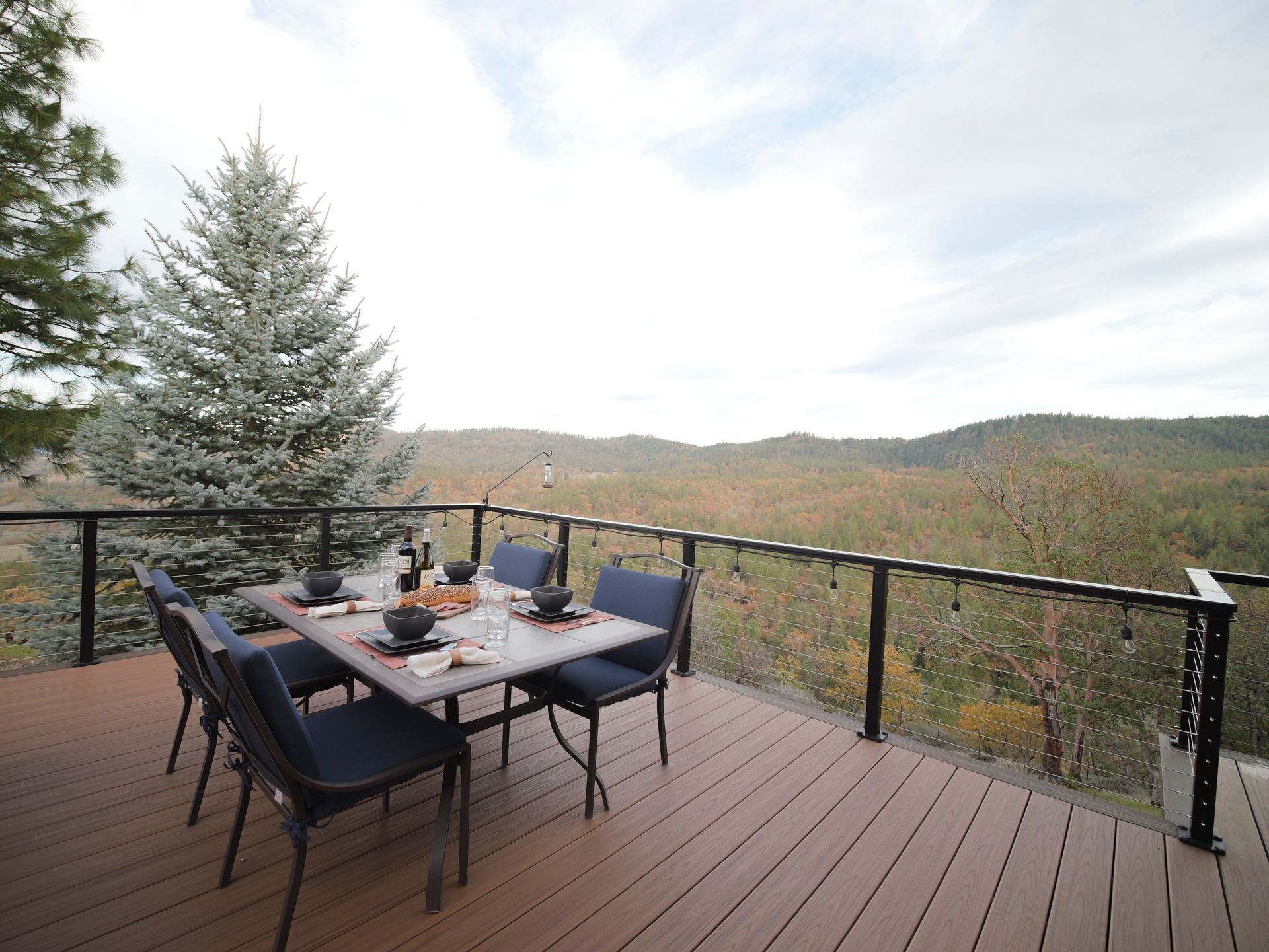 Outdoor dining table set on a deck overlooking a valley. Cloudy sky, evergreen tree.