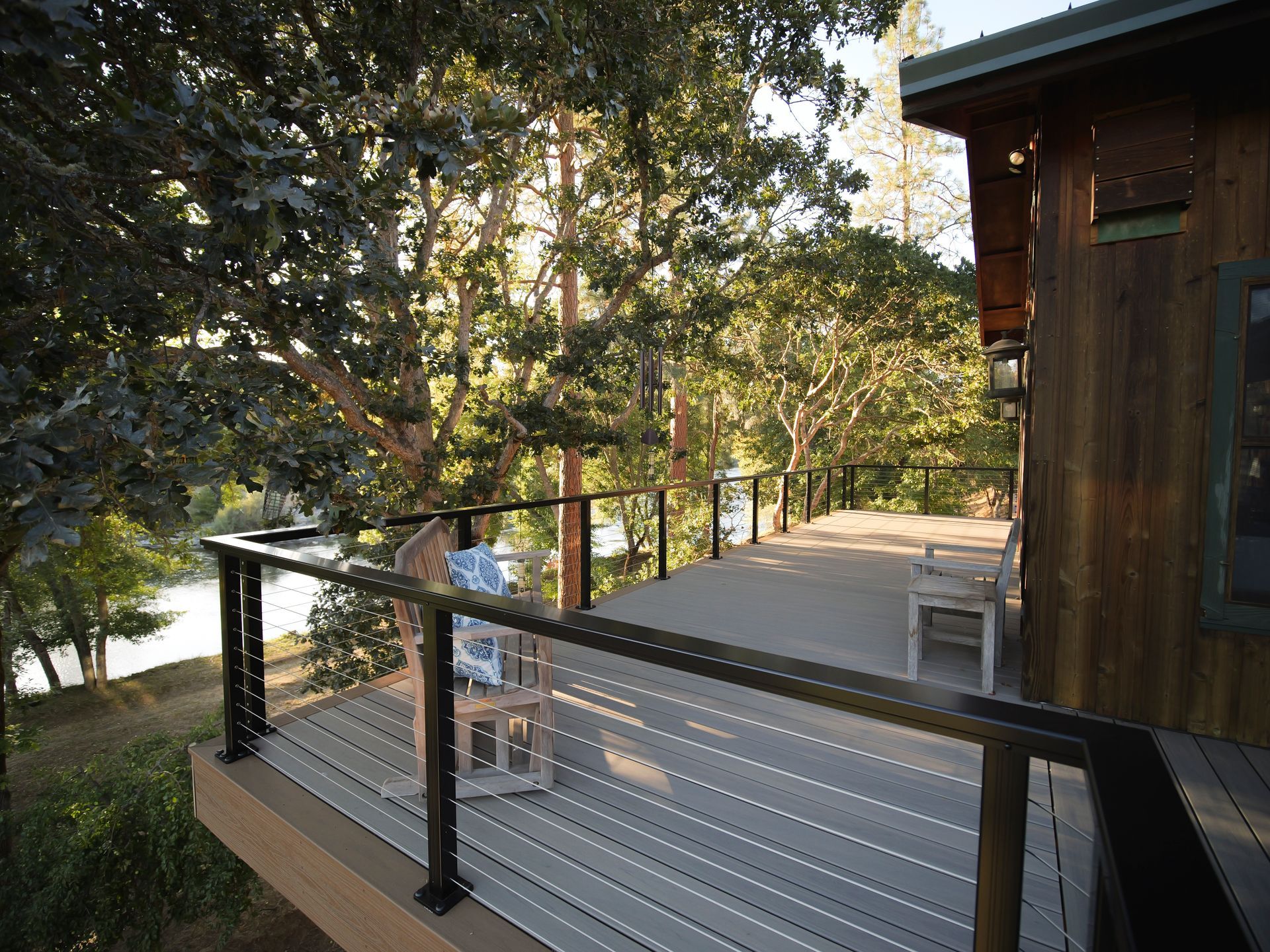 Deck overlooking a river, with a wooden railing and house. Sunlight filters through trees.