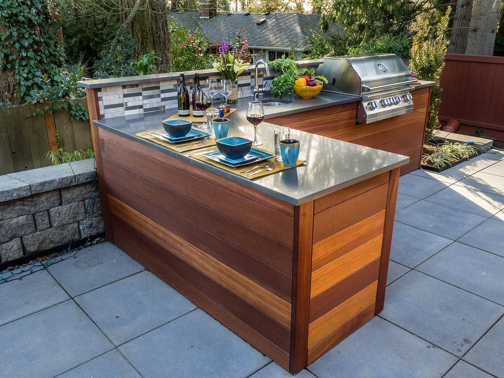 Outdoor kitchen with wooden cabinets, stainless steel countertops, grill, and sink on a patio.