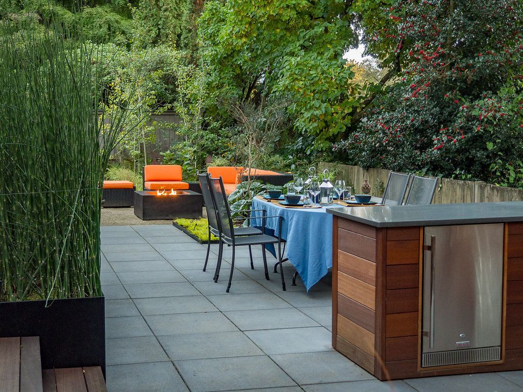 Patio with outdoor kitchen, dining table, seating area, lush greenery, and modern design.