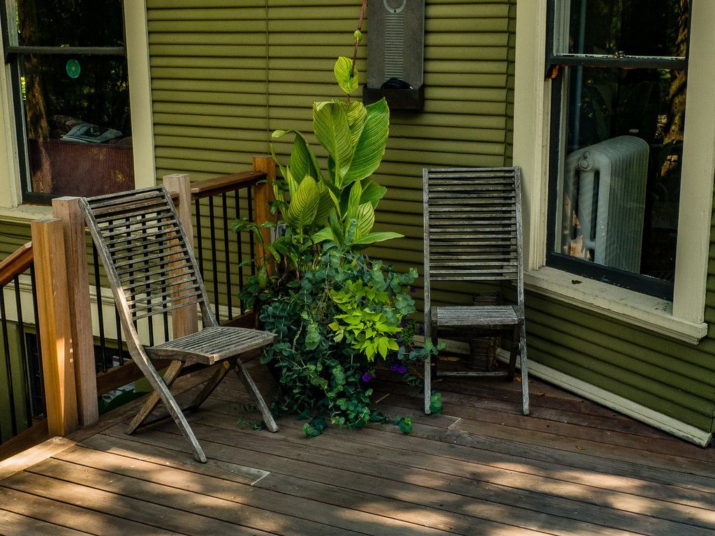 Wooden deck with two chairs, a plant, and two windows on a green house.