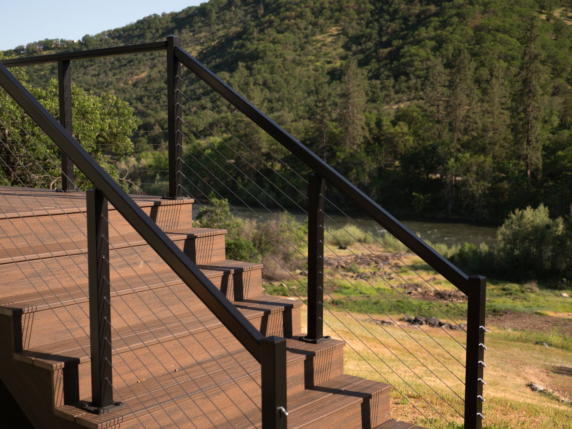 Black metal railing with cable infill on brown deck stairs, overlooking a green hillside and river.