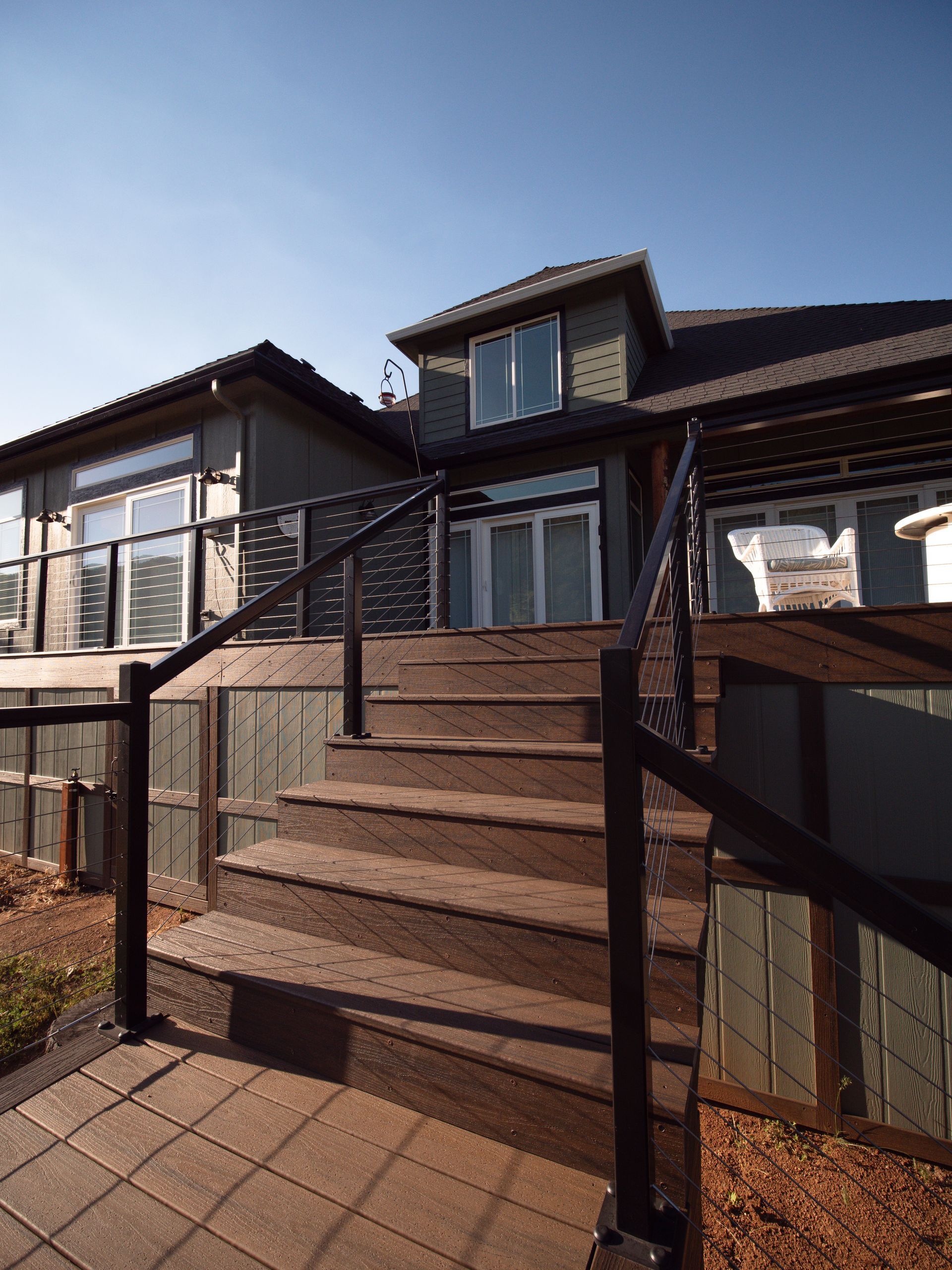 Wooden stairs leading up to a two-story house with black railings and glass panels. Blue sky.