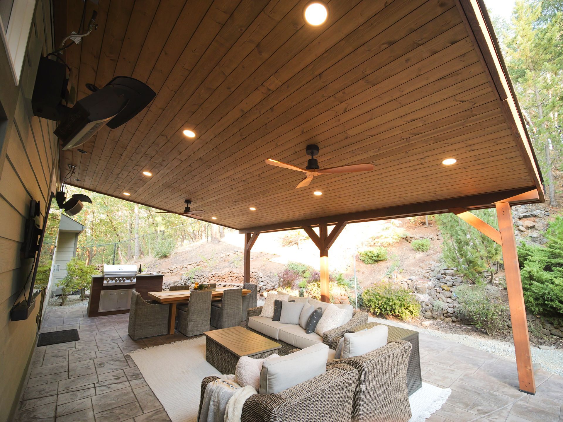 Patio with seating, grill, and wooden ceiling; set in a backyard with forest in the background.