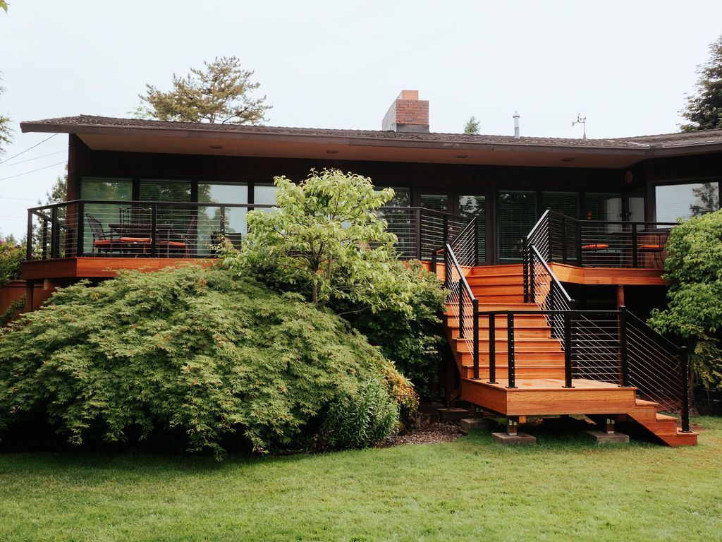Mid-century modern home with wooden deck and stairs, black railings, large windows, and lush greenery.