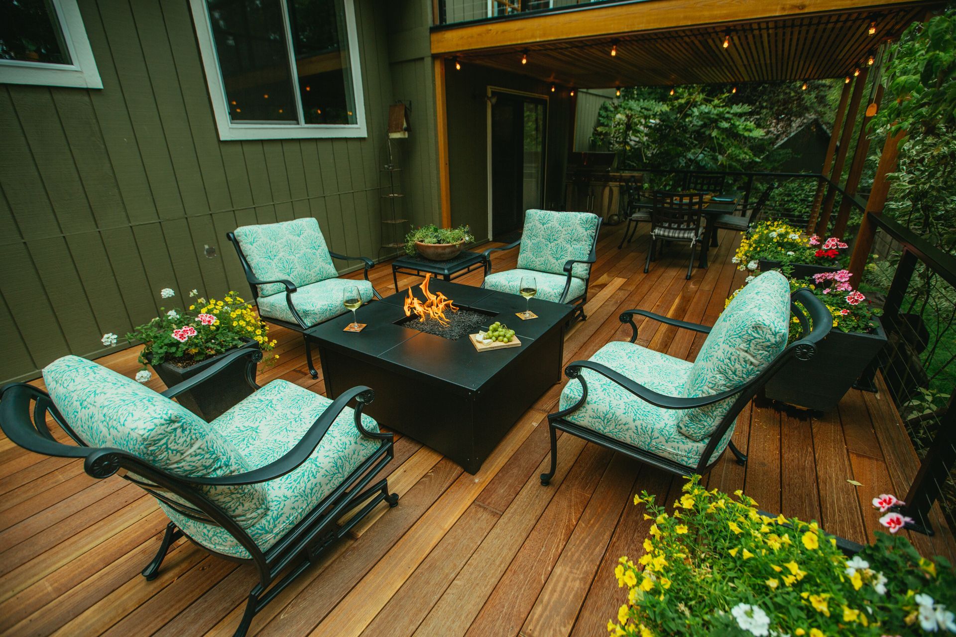 Wooden deck with teal cushioned chairs around a fire pit, surrounded by planters and lush greenery.