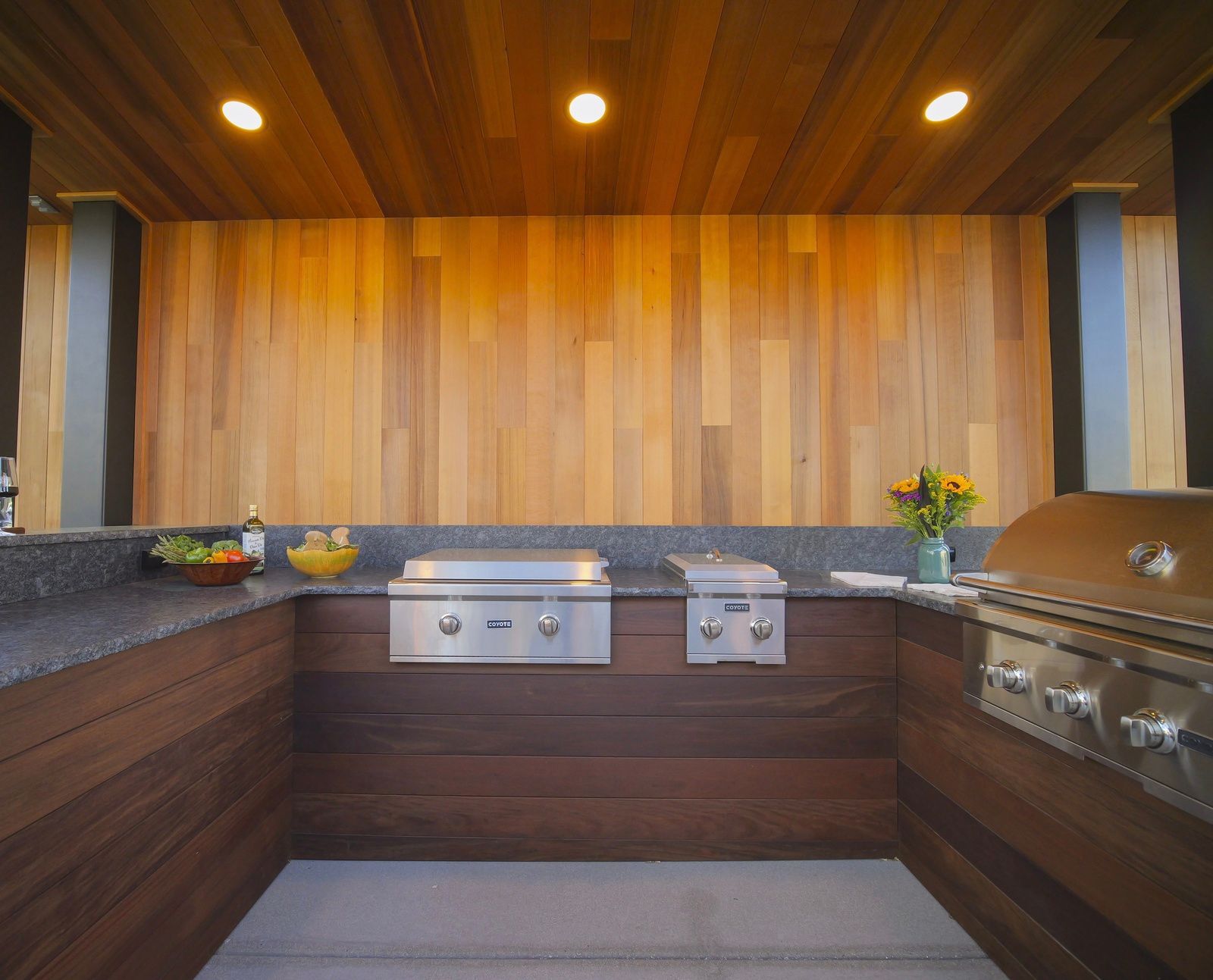 Outdoor kitchen with wooden walls, granite countertop, and stainless steel grills.
