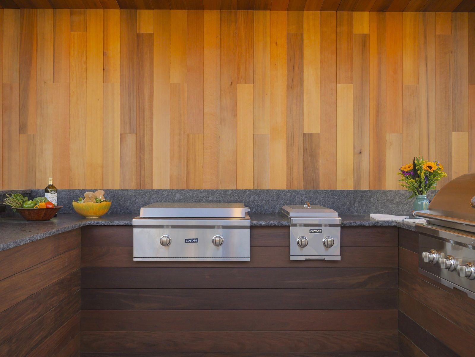 Outdoor kitchen with stainless steel grills, wooden wall, and granite countertop.
