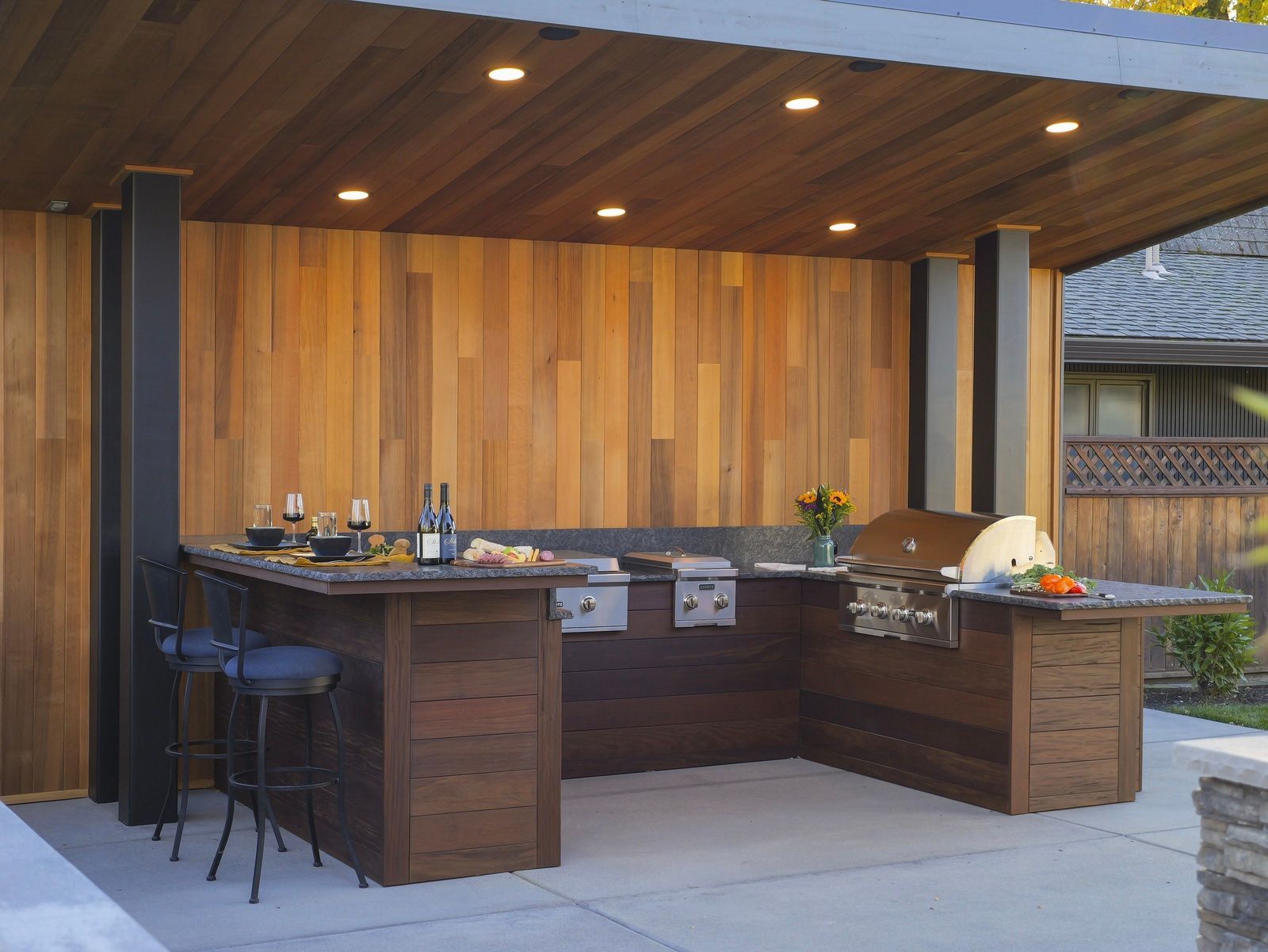 Outdoor kitchen with wood paneling and built-in grill, seating area, and overhead lighting.