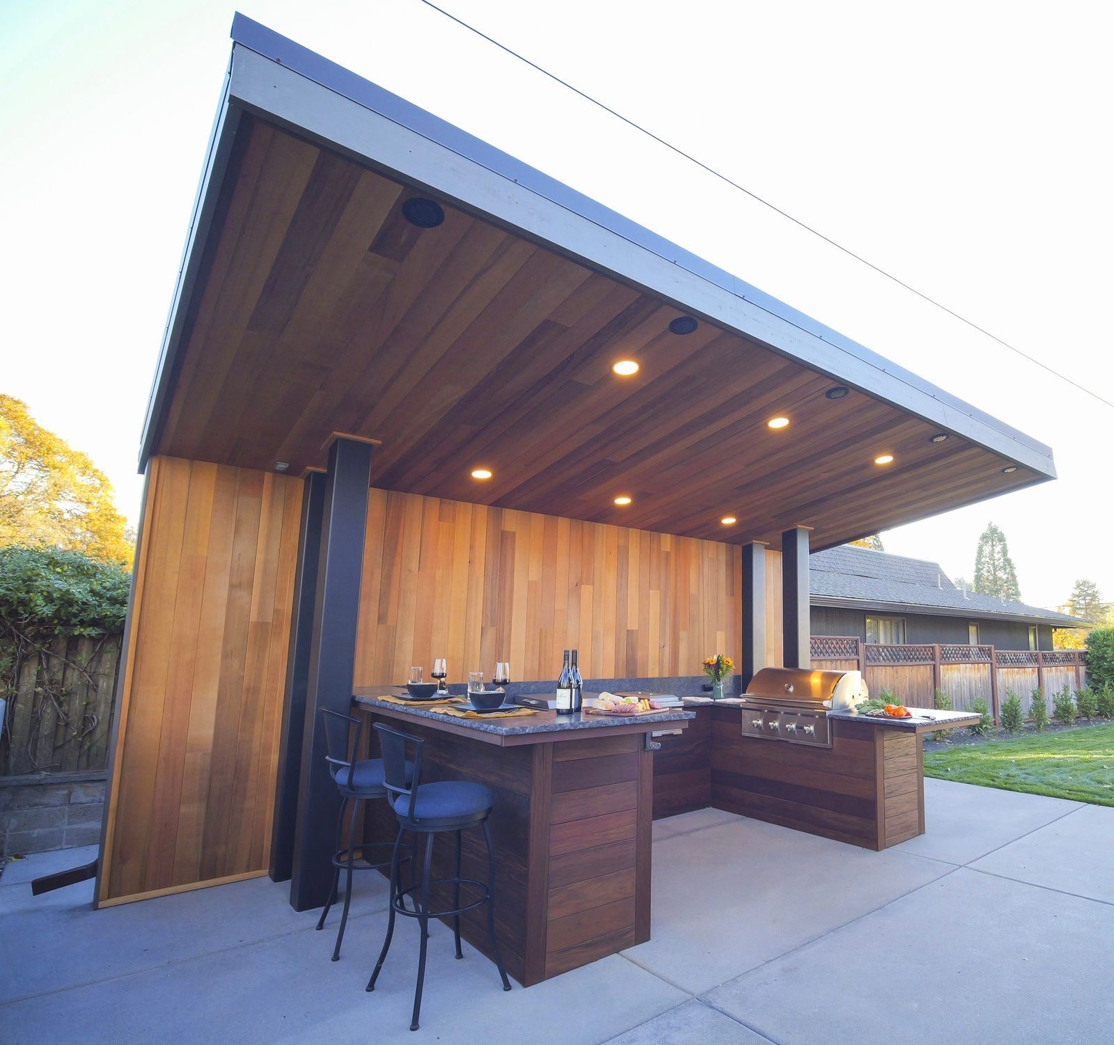 Outdoor kitchen with wood paneling, bar seating, and grill under a modern canopy.