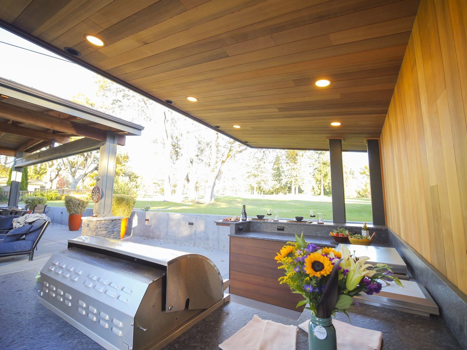 Outdoor kitchen with stainless steel grill, wooden ceiling, countertop, flowers, and view of backyard.