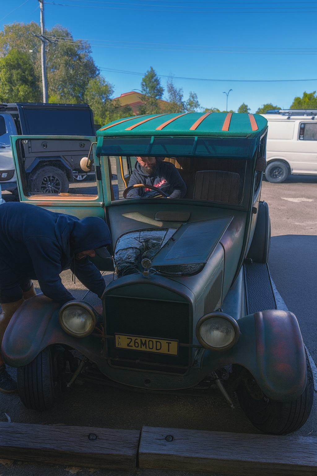 Man working on vintage green car; person in driver's seat. Parked near other vehicles. Sunny day 