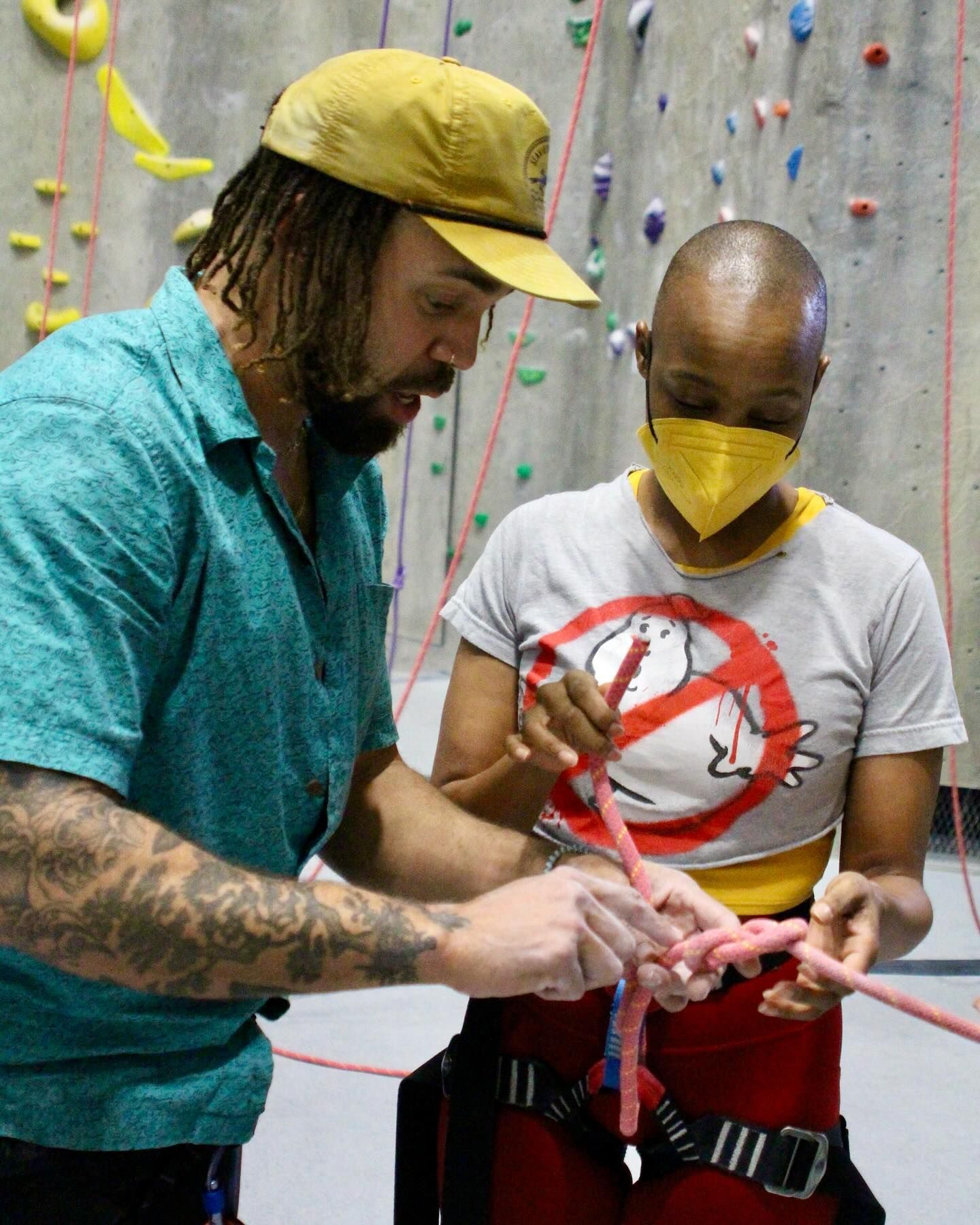Man with tattoos instructs person in harness on knot tying at climbing gym.