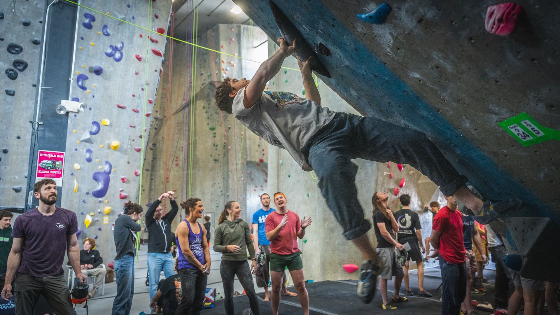 A person bouldering on an indoor climbing wall, surrounded by spectators.