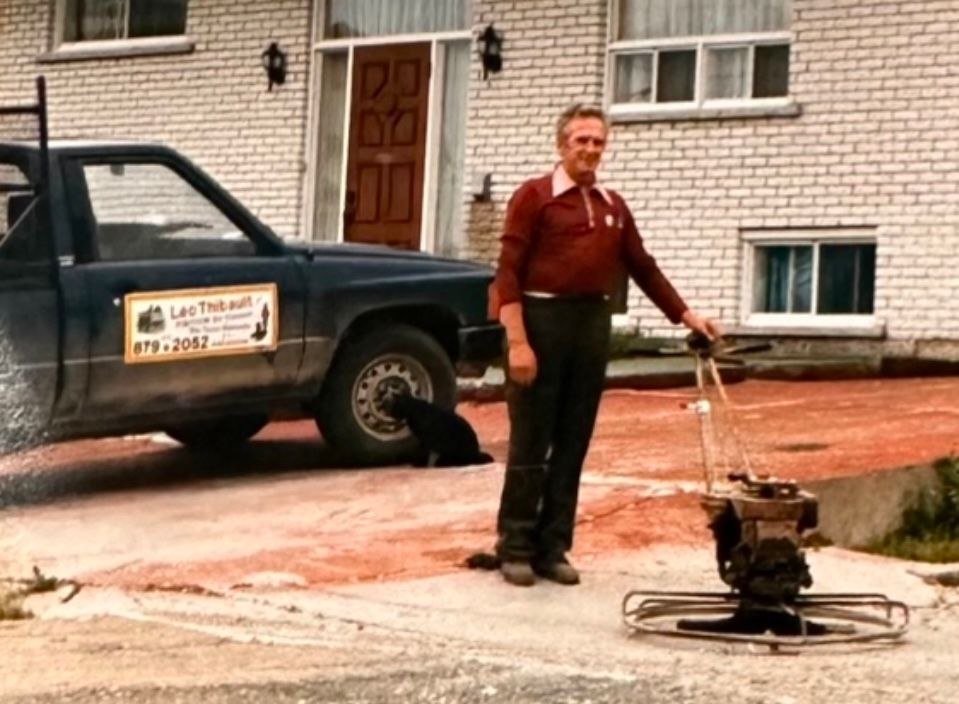 Un homme se tient debout avec une truelle mécanique à côté de son camion devant une maison en briques.