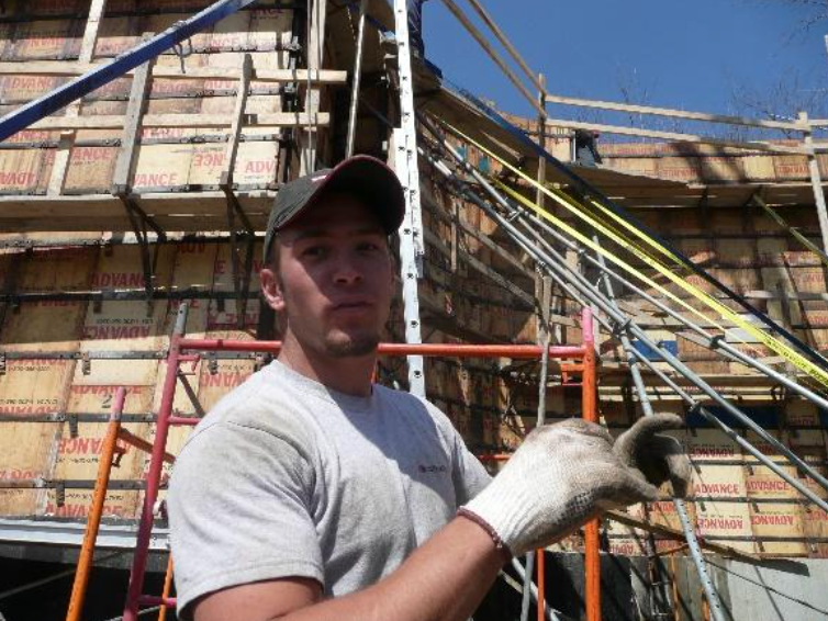 Ouvrier du bâtiment en chemise grise sale et gant, avec un coffrage de bâtiment derrière lui.