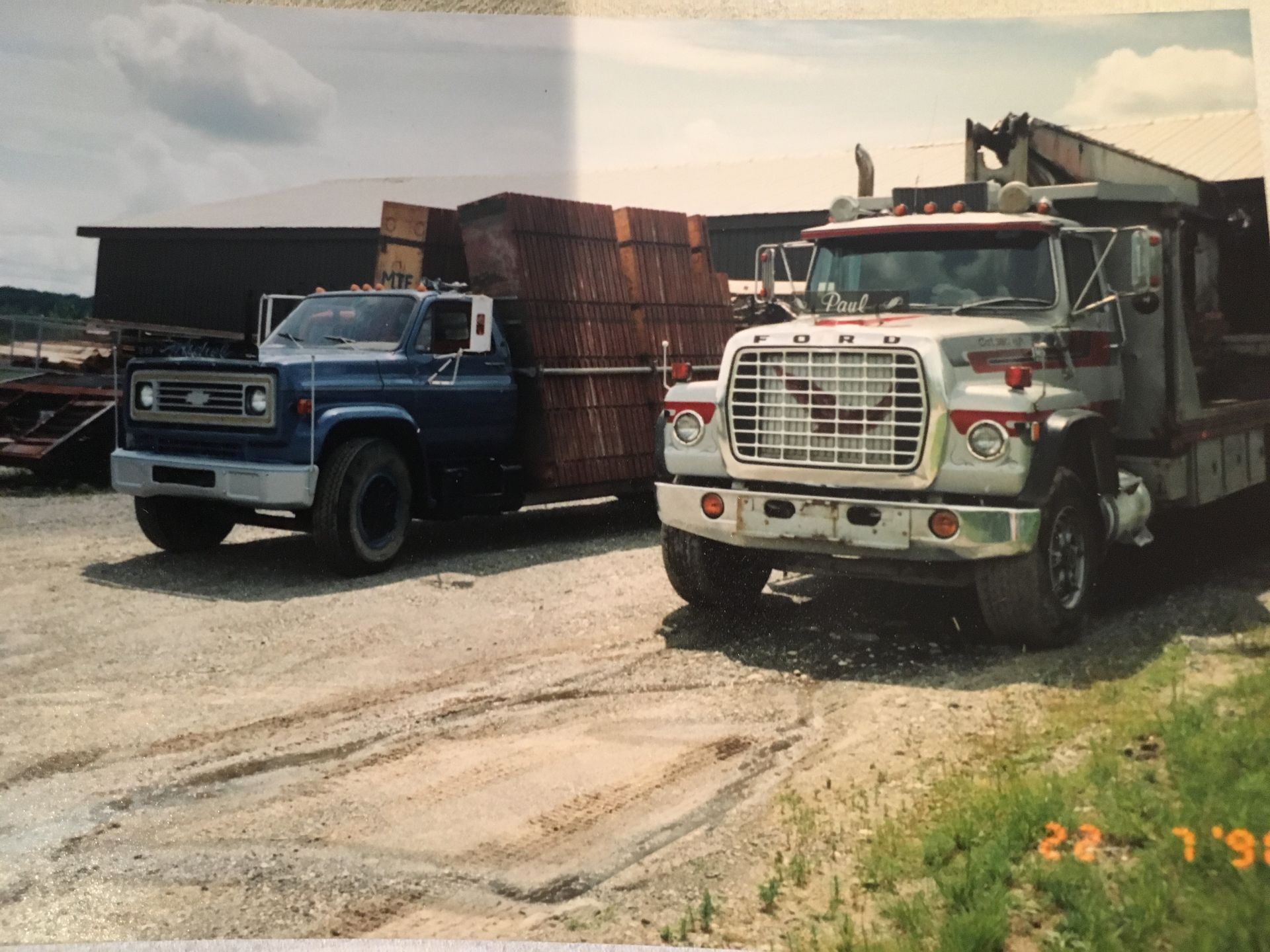 Deux camions chargés de bois sur un terrain en terre battue ; un Chevy bleu et un Ford blanc, datés du 22/1/98.