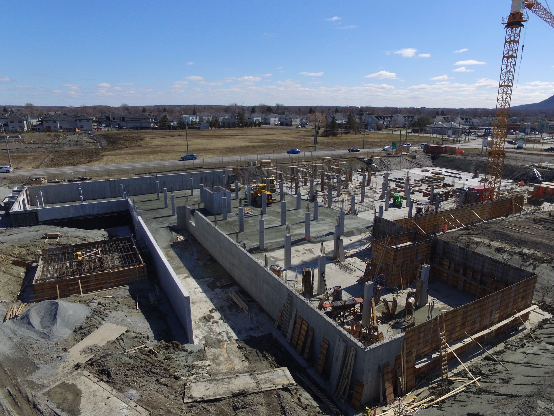 Chantier de construction avec fondations en béton, piliers, murs et grue sous un ciel bleu.
