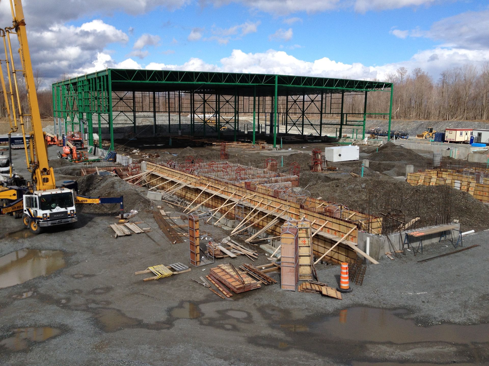 Chantier de construction avec charpente métallique verte, coffrages en bois et camion pompe à béton.