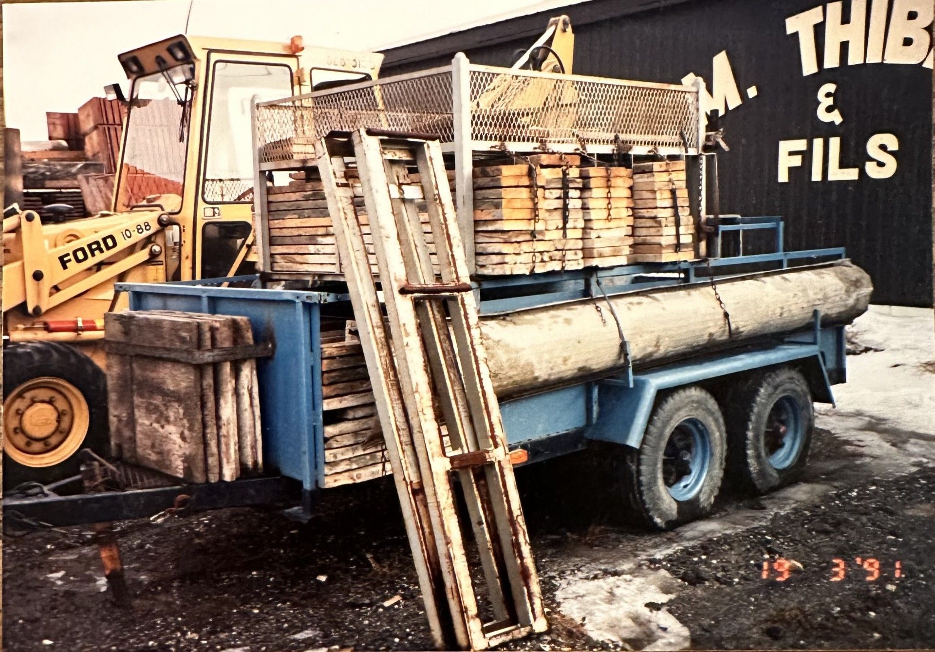 Un tracteur Ford jaune chargeant une remorque avec du bois et des matériaux de construction à l'extérieur d'un bâtiment.