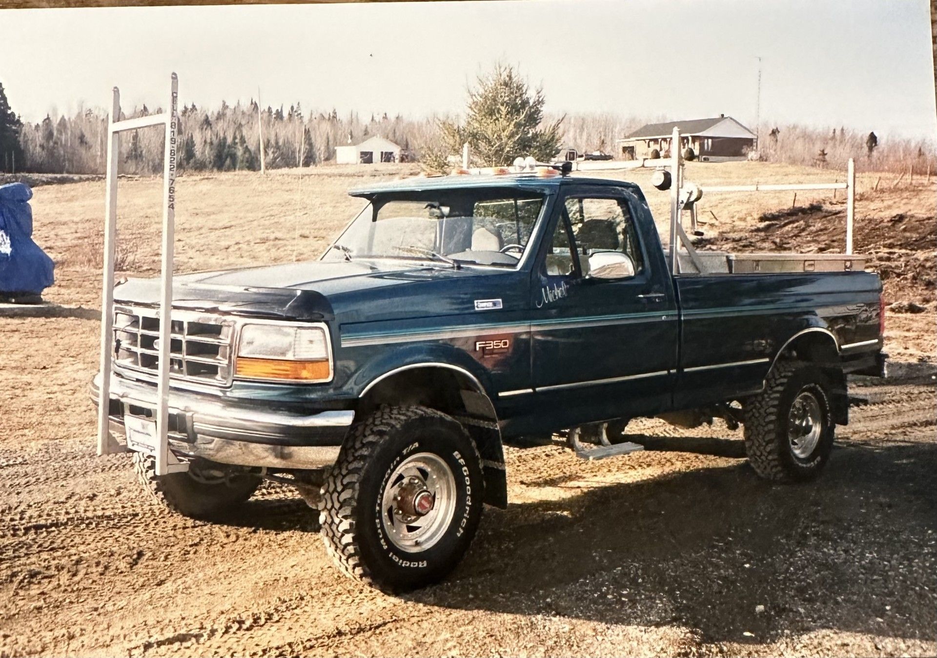 Camionnette Ford bleu foncé avec pneus tout-terrain, sur un chemin de terre, avec des porte-échelles et un arrière-plan rural.
