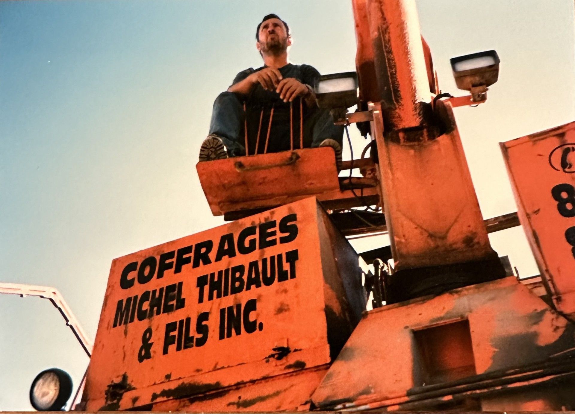 Un homme est assis dans un monte-charge orange, levant les yeux. Un panneau indique « Coffrages Michel Thibault & Fils Inc. ».