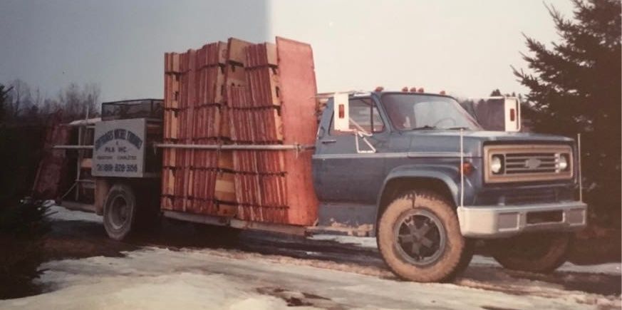 Un camion Chevrolet bleu chargé de planches de bois, garé sur une route enneigée, des arbres en arrière-plan.